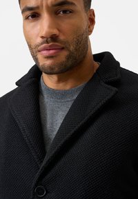 Sort struktureret frakke med et klapkrave, enkelt knaplukning, lag på en grå sweater. Nærbillede af stoffet og detaljen.