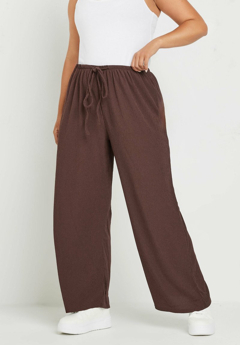 Pantalones marrones de pierna ancha, hechos de una tela texturizada, con una cintura elástica y cordón, y aberturas laterales, combinados con zapatos blancos.