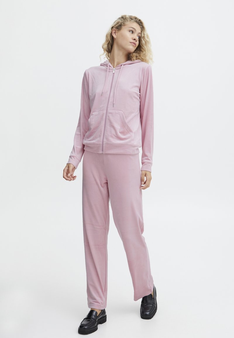 Rosa velour hoodie og bukser sæt med lynlås foran, kengurulommer og lange ærmer, parret med sorte loafers, på en model.