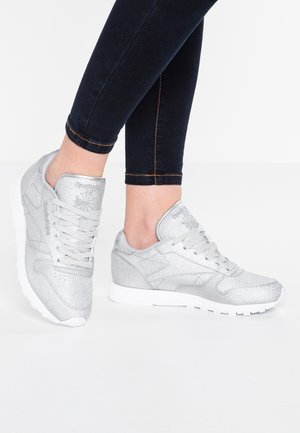 Osoba nosząca srebrne buty sportowe Reebok z białymi podeszwami i czarne dopasowane dżinsy na jednolitym białym tle.