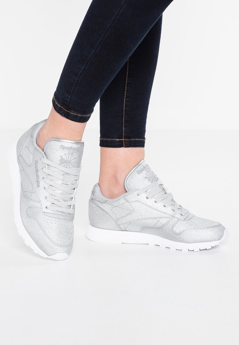 Personne portant des baskets Reebok argentées avec des semelles blanches et un jean noir ajusté sur un fond blanc uni.
