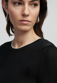 Černý žebrovaný svetr s kulatým výstřihem, který má kontrastní bílý lem. Modelka nosí stříbrné trojúhelníkové náušnice. Hladká textura.