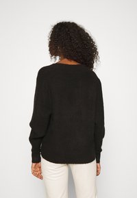 Pull noir en maille côtelée avec manches larges et longues, encolure arrondie et coupe décontractée, associé à un pantalon de couleur claire.