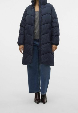 Personne portant un long manteau matelassé bleu marine sur un pull gris, jean large bleu, et bottines à bouts pointus foncées, debout sur un fond uni.