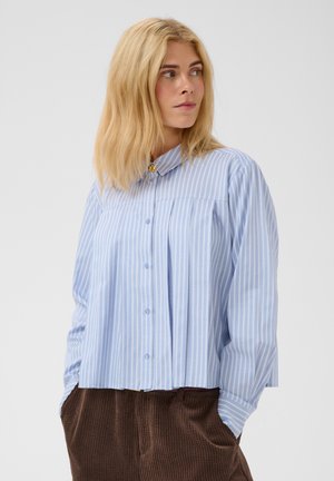 Femme aux cheveux blonds portant une chemise à rayures verticales bleues et blanches à boutons et un pantalon en velours côtelé marron, regardant sur le côté.