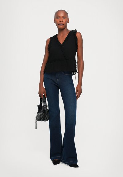 Zwarte gerimpelde peplumtop, gecombineerd met donkere flared jeans. Het model houdt een kleine zwarte mesh tas met kettingdetails vast en draagt puntige hakken.