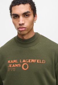 Sudadera verde oliva con cuello alto, presenta el texto en spray naranja "KARL LAGERFELD JEANS" y un diseño de círculo. Textura suave y ajuste casual.