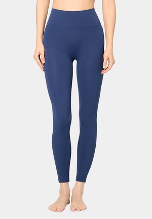 Frau trägt hoch taillierte, marineblaue Leggings in voller Länge und steht barfuß vor einem schlichten hellen Hintergrund.