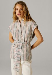 Mujer con cabello rubio ondulado que lleva un chaleco sin mangas a rayas con bordado floral y cinturón de tela, combinado con pantalones blancos, de pie con las manos detrás.