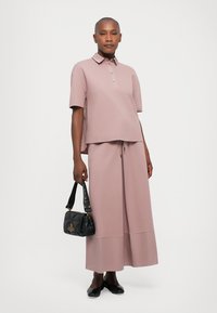Hellrosa übergroßes Polohemd mit kurzen Ärmeln und Knopfleiste, kombiniert mit einem passenden Midirock. Eine schwarze Handtasche und flache Schuhe runden das Outfit ab.