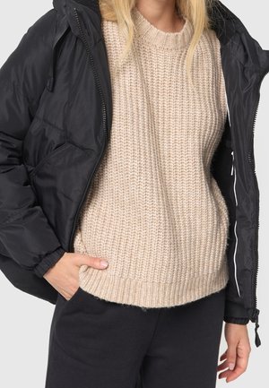 Personne blonde portant un pull en maille beige sous une doudoune noire partiellement zippée, avec une main dans la poche et un pantalon noir.