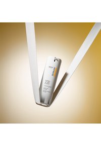Flaske med StriVectin Tighten & Lift Peptight ansigtsløftningsserum holdt oprejst af to hvide elastikbånd mod en gradient beige baggrund.
