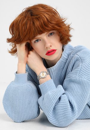 Montre-bracelet en argent avec un cadran rond et des accents en cristal, reposant sur un pull en maille bleu clair avec des poignets côtelés.