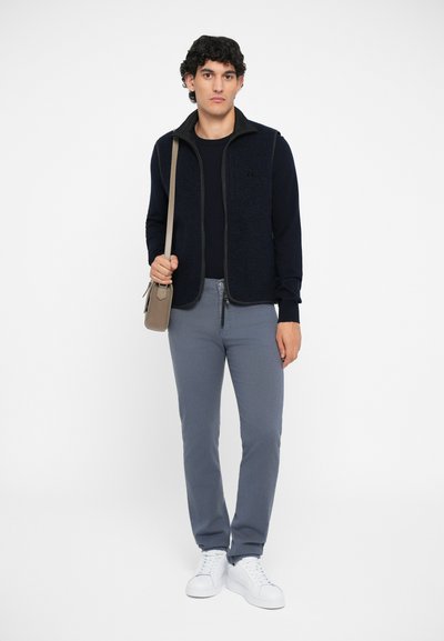 Navy fleece vest over a navy long-sleeve shirt, gray slim-fit pants, and white sneakers. Brown shoulder bag with a strap.