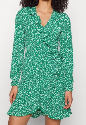 Kvinde iført en grøn slå-om-kjole med hvidt blomsterprint og flæsedetaljer langs udskæringen og nederdelen.