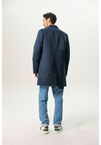 Marineblauer langer Mantel mit taillierter Passform, Rückenschlitz und glatter Textur, kombiniert mit hellblauen Jeans und mehrfarbigen Turnschuhen.