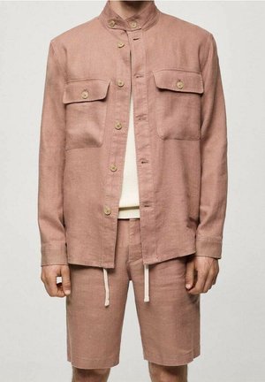 Homme portant une veste boutonnée marron clair avec deux poches avant et un short assorti avec des cordons blancs ainsi qu'une chemise crème en dessous.