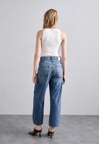 Kvinna med blont hår står vänd mot en vanlig vägg, klädd i en vit ärmlös topp, blå jeans och bruna klackskor på en grå golv.