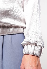 Detailní záběr ruky oblečené ve lesklé stříbrné blůze s dlouhým rukávem a nařasenými manžetami, doplněné vysokým modrým sukní.