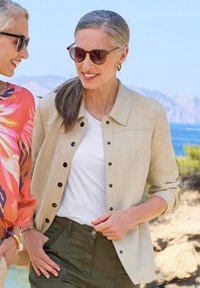 Lichtbeige suede shirtjack met zwarte drukknoopjes, gedragen over een wit T-shirt; gecombineerd met donkergroene broeken. De achtergrond toont een natuurlijke landschap.