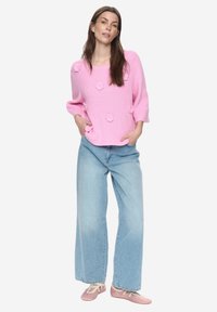 Růžový pletený svetr s květinovými motivy, širokými rukávy, doplněný světle modrými džínami se širokými nohavicemi a jemnými růžovými balerínkami.