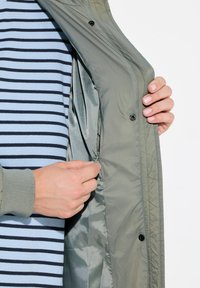 Lichte olijfgroene jas met open knopen, die een gladde voering onthult. Gestreept blauw-zwart shirt eronder, met lange mouwen.