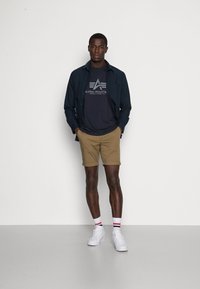 Herrestil med en marineblå skjorte med knapper, mørkegrå t-shirt med grafik, beige shorts og hvide sneakers med røde og blå stribede sokker.