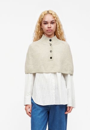 Woman with long curly blonde hair wearing a beige knit buttoned capelet over a white striped shirt and blue jeans against a white background.