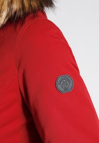 Rød jakke med en glat, mat overflade. Har en krave af kunstpels og et sort logo-patch på den højre ærme.