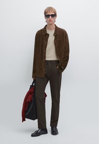 Camicia in suede marrone, maglione crema, pantaloni marroni scuri, scarpe nere e occhiali da sole. Tiene una borsa con un oggetto rosso. Design semplice e moderno.