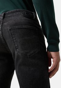 Černé džíny z denimu s hladkou texturou, vybavené zadní kapsou a kulatou černou nášivkou s logem na pase. Design je slim fit.