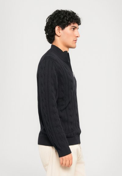 Νεαρός άνδρας με καστανά σγουρά μαλλιά, που φοράει ένα μαύρο ζακάρ φούτερ με φερμουάρ και ανοιχτό μπεζ παντελόνι, στέκεται σε προφίλ μπροστά από έναν απλό φόντο.