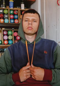 Der Kapuzenpullover hat ein Farbblockdesign mit dunkelgrünen, marineblauen und burgunderroten Abschnitten, akzentuiert durch eine orangefarbene Kordel und ein kleines Elefantenlogo.