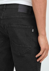 Mustad denim püksid sujuva tekstuuriga, kahel tagaosal taskud ja must nahast silt vöökoht. Nähtav detailne õmblemine.