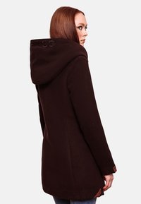 Manteau à capuche marron avec une texture douce, au design allongé, et des détails brodés sur la capuche. Présente un accent de logo au dos.