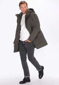 Olivengrøn polstret parka-jakke med en grå hætteforing, frontlynlås og knapstolpe; parret med mørke jeans og sorte støvler.