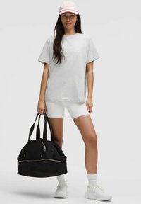 T-shirt gris, shorts de vélo blancs et baskets blanches. Casquette rose, sac de sport noir avec des zippers et de larges sangles noires.