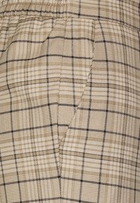 Beige ternet stof med et gittermønster og tynde sorte linjer. Teksturen virker vævet med synlige sømme og lomme-detaljer.