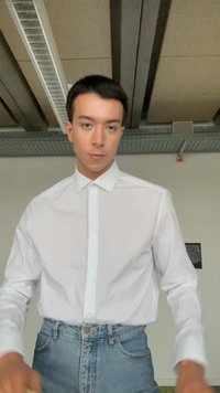 Jeune homme portant une chemise blanche à boutons et un jean bleu, debout à l'intérieur contre un mur clair avec des poutres apparentes au plafond.