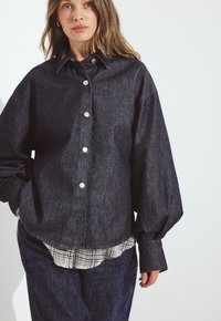 Tmavé denimová oversized košeľa s golierom, striebornými gombíkmi a balónovými rukávmi, nasadená na károvanú košeľu.