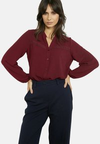 Burgundia burgundi toonides pikkade varrukatega pitsdetailidega pluus õlgadel. Sobitatud kõrge vöökohaga tumesiniste pükstega. Sile kangapind.