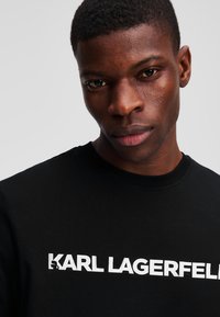 Juodas apvalaus kaklo marškinėliai su balto spaudiniu "KARL LAGERFELD", pasižymintys lygiu paviršiumi ir standartiniu kirpimu.