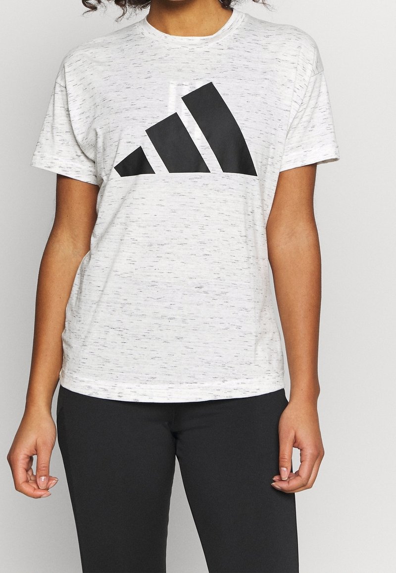 Personne portant une chemise blanche à manches courtes avec un grand logo géométrique noir sur la poitrine et un pantalon noir, debout devant un fond uni.
