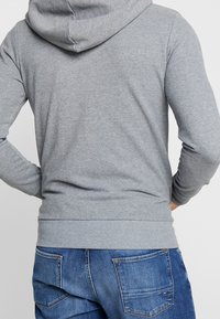Sudadera con capucha gris, con puños y dobladillo acanalados, vista desde la parte de atrás. Combinada con jeans de mezclilla azul, que presentan una textura desgastada.