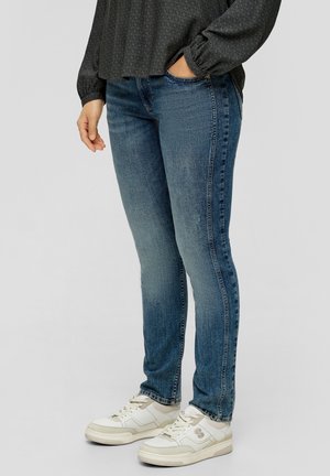 Schmale Jeans aus mittelblauem Denim, mit glatter Textur, sanften Verwaschungen und klassischem Fünf-Taschen-Design. Getragen mit weißen Sneakern.