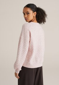 Világos rózsaszín kötött pulóver, texturált cikk-cakk mintával, kerek nyakkivágással, bordázott mandzsettákkal és szegéllyel. Sötét nadrág fölött viselve.