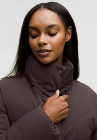 Doudoune marron avec un col haut, tissu texturé et bouton-pression. Elle présente une poche avant et est portée avec une main tenant le col.