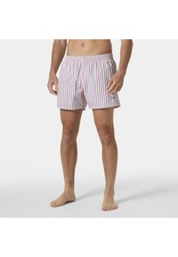 Hombre vestido con bañador blanco con rayas verticales rojas y azules, de pie descalzo sobre un fondo blanco.