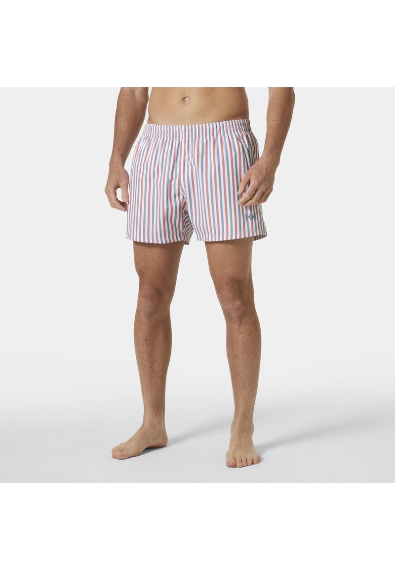 Hombre vestido con bañador blanco con rayas verticales rojas y azules, de pie descalzo sobre un fondo blanco.