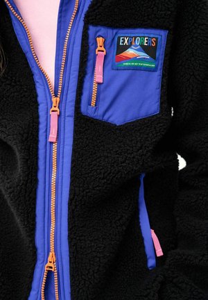 Czarna polarowa kurtka z niebieską kieszenią na piersi i wykończeniem, pomarańczowymi zamkami z różowymi suwakami oraz kolorową naszywką „Explorers” z motywem gór.